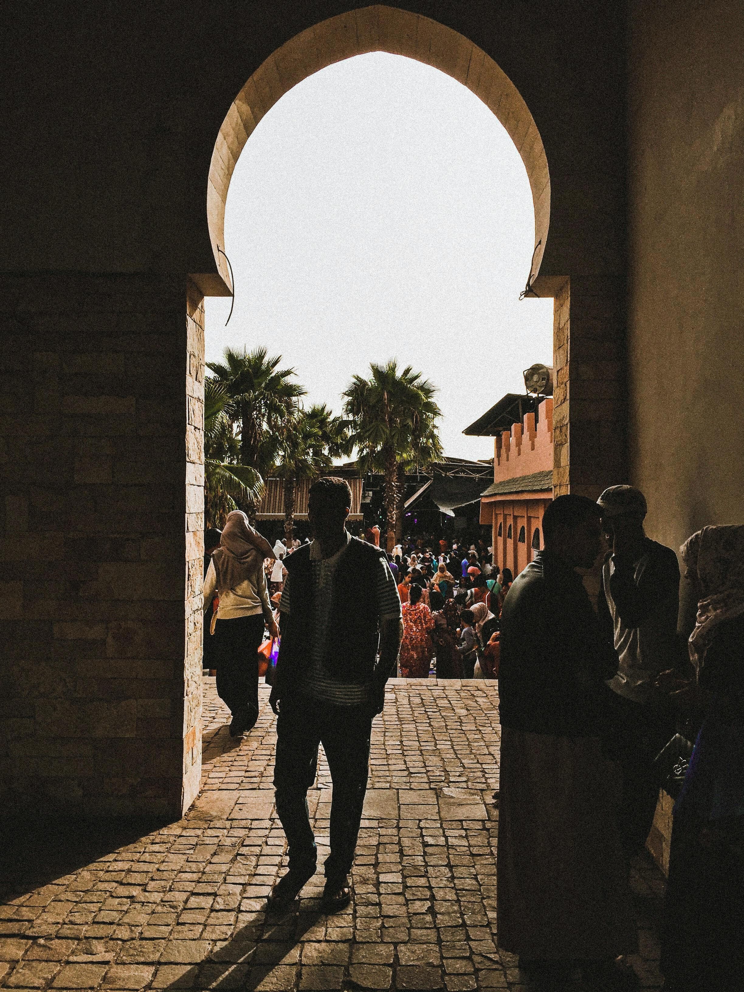 An arched doorway leading to a crowded square with palm trees and buildings in the background.