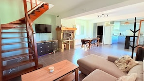 A living room with a wooden staircase, a TV, a fireplace, a dining area, and a kitchen.