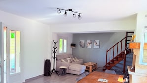 Un salon comprenant un canapé, une table basse et une table à manger. On y trouve également un porte-manteau, un escalier et une porte donnant sur l’extérieur.