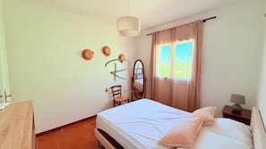 A bedroom with a bed, a chair, a mirror, a hanging light, and a window with curtains.