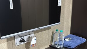 A hotel room with a flat-screen TV mounted on the wall, a dark wooden console table, a water bottle, a remote control, and a blue towel. - Hotel Shri Ji Palace (Rajnagar)