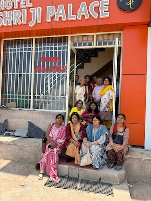 Un grupo de personas sentadas frente a un hotel llamado 'Hotel Shri Ji Palace', con un letrero que indica que hay habitaciones disponibles.