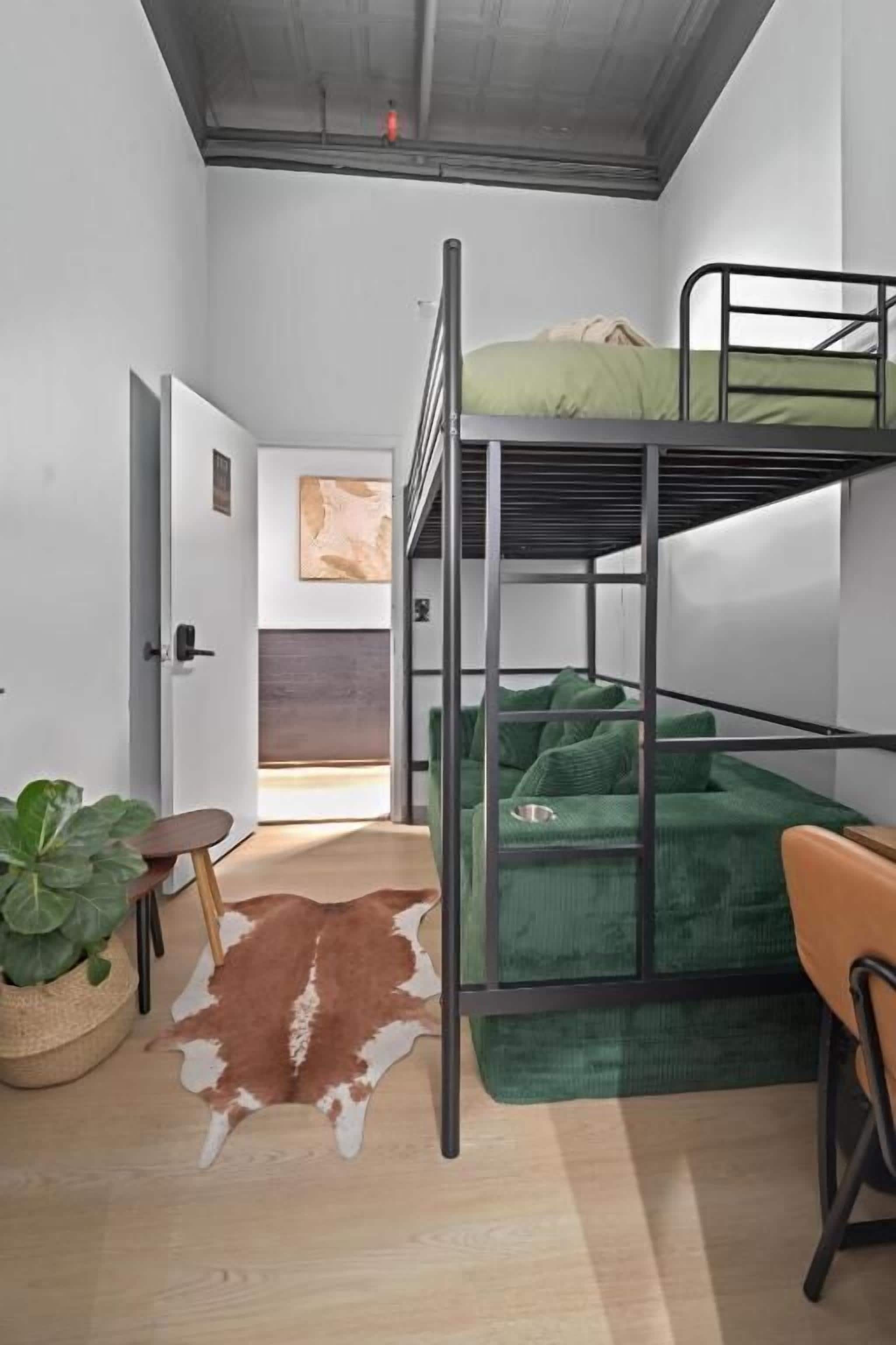 A bunk bed with green bedding, a wooden floor, a cowhide rug, a small table with a plant, and a painting on the wall.