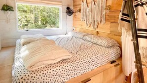 A wooden cabin bedroom with a bed, a window, a wall-mounted fan, and macrame decorations.