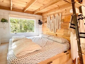 Une chambre dans une cabane en bois, avec un lit, une fenêtre, un ventilateur fixé au mur et des décorations en macramé.
