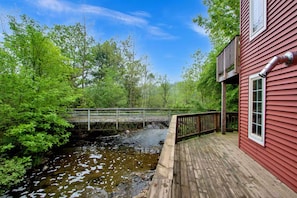 A wooden deck adjacent to a red building with a metal railing and a small bridge over a stream. - 2-bedroom mill in adorable Wautoma with AC and charm (Wautoma)