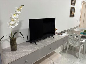 A modern living room with a flat-screen TV, a vase with white flowers, and a dining area with a table and chairs. - Ravello Residential AP04 Foz do Iguaçu (Foz do Iguaçu)
