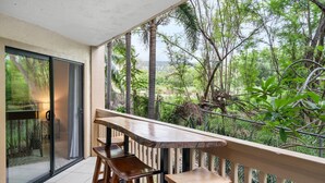 A balcony with a wooden table and two chairs, overlooking a lush green landscape with trees and a view of the sea. - Royal Kahili B204 AC Across From Lymans/banyans (Kailua-Kona)