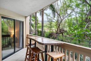 Ein Balkon mit Holztisch und zwei Stühlen, der auf eine üppige, grüne Landschaft mit Bäumen und Meerblick blickt.