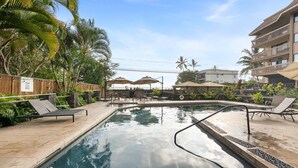 A swimming pool with lounge chairs and umbrellas, surrounded by palm trees and a wooden fence. - Royal Kahili B204 AC Across From Lymans/banyans (Kailua-Kona)