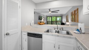 A modern kitchen with a dishwasher, double sink, ceiling fan, and a view of greenery outside. - Royal Kahili B204 AC Across From Lymans/banyans (Kailua-Kona)