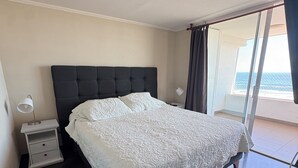 Een slaapkamer met een groot bed, een nachtkastje, een lamp en uitzicht op de zee door een groot raam.