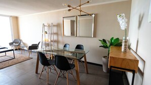 Een moderne eethoek met een glazen tafel, zwarte stoelen, een houten bijzettafeltje met een plant en een decoratieve spiegel aan de muur.