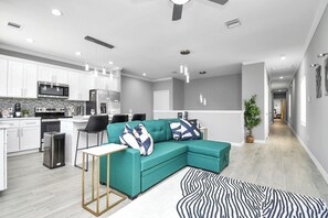 Een moderne keuken met witte kasten, een magnetron en een koelkast. Er staat een blauwgroene bank met gestreepte kussens, een zwarte stoel en een klein tafeltje. Een vloerkleed met zebraprint ligt op de grond.