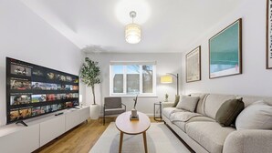 A modern living room with a flat-screen TV, a round coffee table, a sofa, a window, and a floor lamp. - 2BR • New Listing Discount • Free Parking (West Sussex)