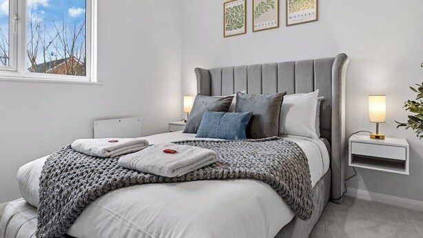 A modern bedroom with a grey upholstered bed, white bedding, and a bedside table with a lamp. - 2BR • New Listing Discount • Free Parking (West Sussex)