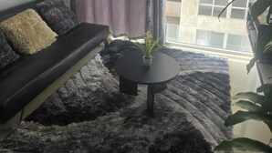 Ett vardagsrum med en svart soffa, ett runt soffbord och ett stort fönster med gardiner.