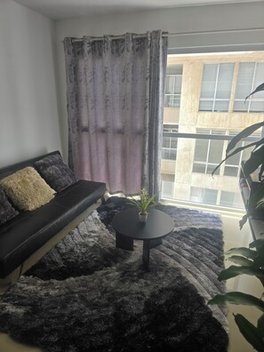 Ein Wohnzimmer mit einem schwarzen Sofa, einem runden Couchtisch und einem großen Fenster mit Vorhängen.
