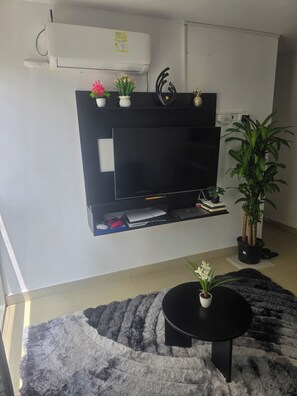 Ein Wohnzimmer mit Wand-Fernseher, einem schwarzen Couchtisch, einer Topfpflanze und einem schwarzen Teppich.