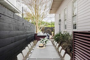 A covered outdoor dining area with a long table, chairs, and a grill. - Stylish Designer Home  | Shared Heated Pool + Spa (New Orleans)