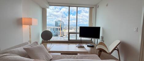 A modern hotel room with a large window, a flat-screen TV, a sofa, a bedside table, and a lamp.