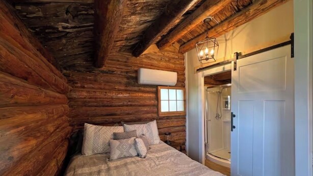 A log cabin bedroom with a bed, pillows, a window, and a door leading to a shower area. - Cozy Rustic Cabin in the City (Richfield)