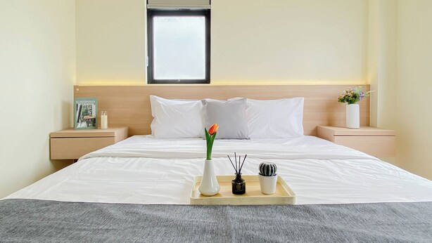 A hotel room with a bed, bedside tables, a nightstand, a window, and a tray with a vase and a plant. - Neat 3Br For 4 Pax At Meikarta District 2 Apartment (Cikarang)