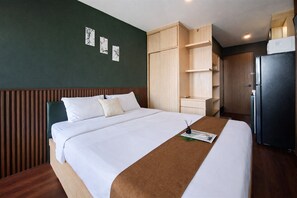 A hotel room with a bed, wooden headboard, a wardrobe, a refrigerator, and wall art. - Green Urban Style Studio Room At Apartment Sudimara Forestwalk (South Tangerang)