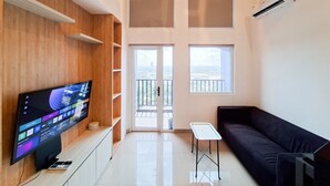 A modern living room with a flat-screen TV, a dark sofa, a small table, and a balcony with a city view. - Minimalist Studio Loft Apartment At Kingland Avenue (South Tangerang)