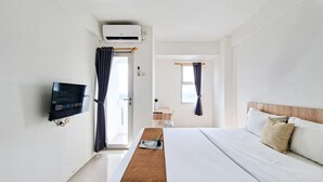 A hotel room with a bed, a TV mounted on the wall, a window with curtains, and an air conditioning unit. - Cozy Studio Apartment At Urbantown Serpong (South Tangerang)