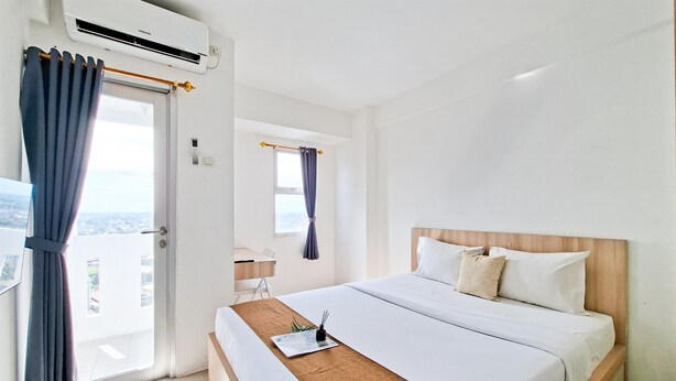 A hotel room with a bed, a window with curtains, an air conditioning unit, and a view of the outside. - Cozy Studio Apartment At Urbantown Serpong (South Tangerang)