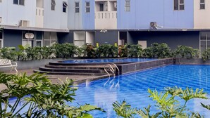 A swimming pool with steps leading into it, surrounded by greenery and a multi-story building with balconies. - Cozy Studio Apartment At Urbantown Serpong (South Tangerang)