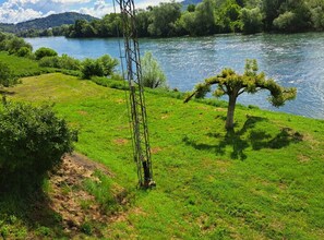 Ein Fluss mit einer grasbewachsenen Uferböschung, einem Baum und einer Strommast-Konstruktion.