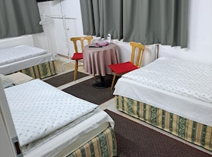 Una habitación de hotel con dos camas individuales, una mesita con una silla, un radiador y una ventana con cortinas.