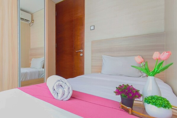 A hotel room with a wooden door, a bed with white bedding, a pink bedspread, a rolled towel, and two potted plants.
