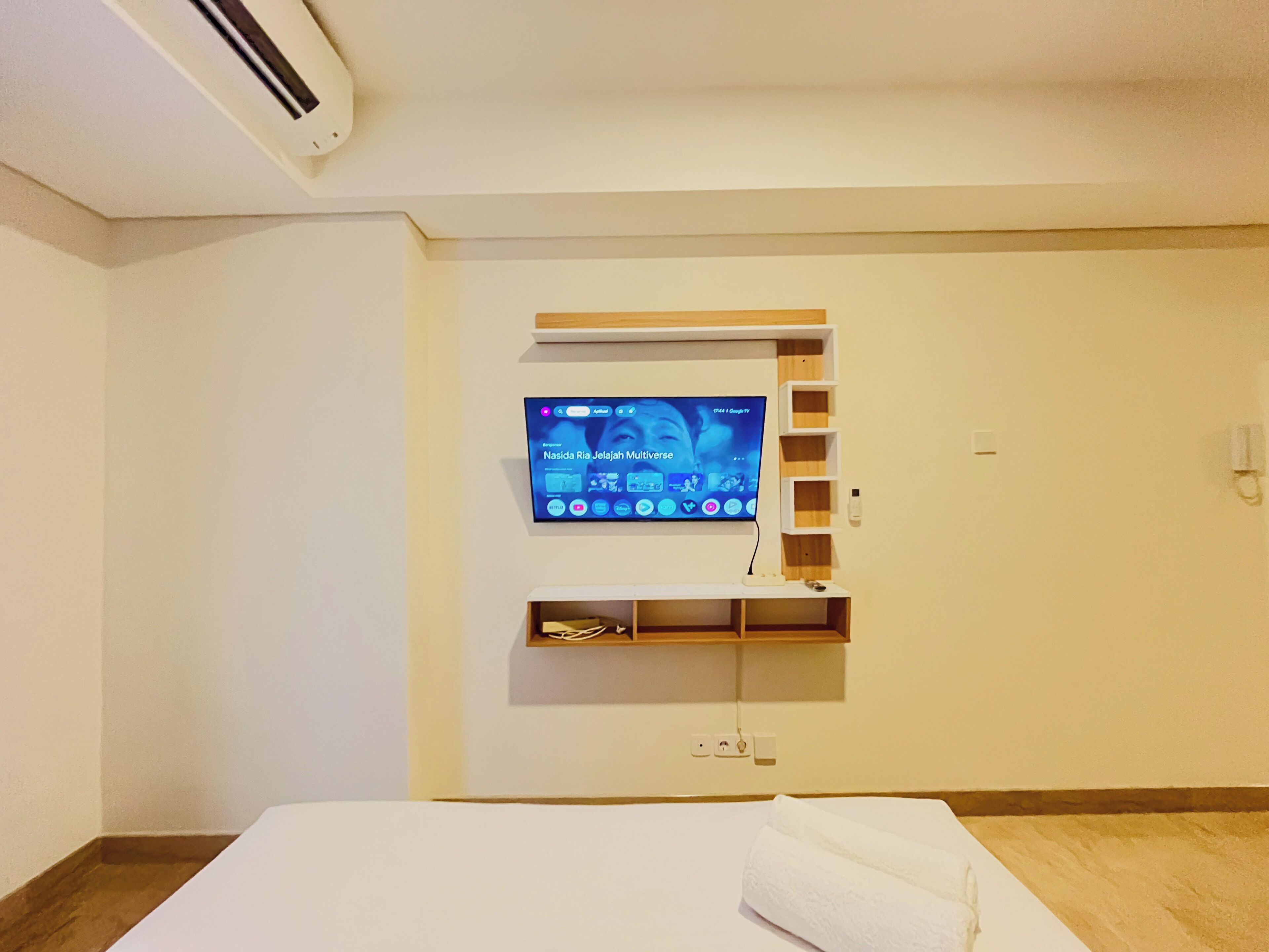 Una camera d'albergo con una TV a schermo piatto montata sulla parete, uno scaffale in legno sottostante e un letto con copripiumino bianco.