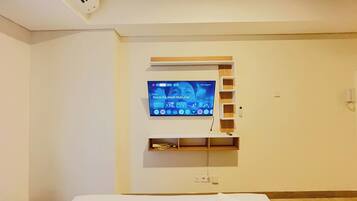 Una camera d'albergo con una TV a schermo piatto montata sulla parete, uno scaffale in legno sottostante e un letto con copripiumino bianco.