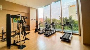 A gym with cardio machines and weight equipment, large windows with a view of greenery and a building. - Chic And Warm Studio With Super King Bed At Capitol Suites Apartment (Jakarta)