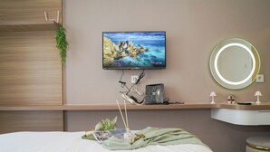A hotel room with a bed, a TV mounted on the wall, a bedside table with a lamp, a round mirror, and a small plant. - Chic Brown Touch Studio At Apartment Collins Boulevard (Tangerang)