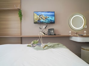 A hotel room with a bed, a TV mounted on the wall, a bedside table with a lamp, a round mirror, and a small plant. - Chic Brown Touch Studio At Apartment Collins Boulevard (Tangerang)