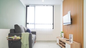 A modern living room with a grey sofa, a small wooden side table, a TV mounted on a wooden wall, and a large window with sheer curtains. - Bright And Relaxing 2Br At Collins Boulevard Apartment (Tangerang)