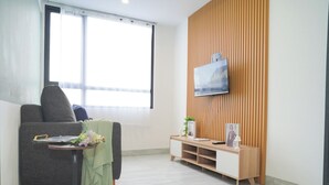 A living room with a sofa, a small table with a plant, a TV on a wooden wall, and a window with blinds. - Bright And Relaxing 2Br At Collins Boulevard Apartment (Tangerang)