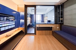 Quarto de hotel moderno com piso de madeira, parede azul, televisão grande, escrivaninha, um banco e vista para o quarto vizinho.