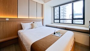 A hotel room with a large bed, built-in headboard, a window with a view of buildings, and a bedside table with a magazine. - Best Deal Studio Apartment Collins Boulevard (Tangerang)