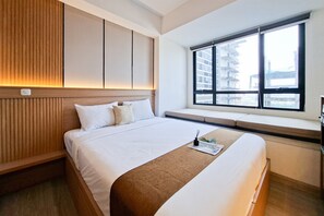 Ein Hotelzimmer mit einem großen Bett, einer festen Kopfstütze, einem Fenster mit Blick auf Gebäude und einem Nachttisch mit einer Zeitschrift.