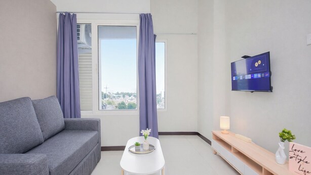A modern living room with a grey sofa, a small white table with a tray, a TV mounted on the wall, and a window with curtains. - Stunning Studio Loft At Nifarro Park Apartment (Jakarta)