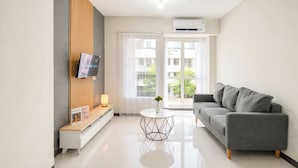 Ein modernes Wohnzimmer mit grauem Sofa, einem kleinen weißen Couchtisch, einem an einer Holzplatte montierten Fernseher und einem großen Fenster mit Blick auf Gebäude und Bäume.