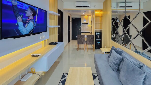 A modern living room with a flat-screen TV, a sofa, a coffee table, and a dining area with chairs and a table. - Neat Warm Wooden Tone 1Br At Patraland Amarta Apartment (Ngaglik)