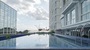 A modern hotel with a swimming pool, large windows, and a clear sky. - Neat Warm Wooden Tone 1Br At Patraland Amarta Apartment (Ngaglik)