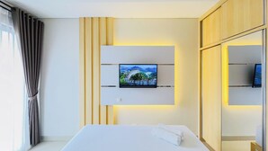 A modern hotel room with a bed, a flat-screen TV mounted on the wall, and a large mirror. - Neat Warm Wooden Tone 1Br At Patraland Amarta Apartment (Ngaglik)